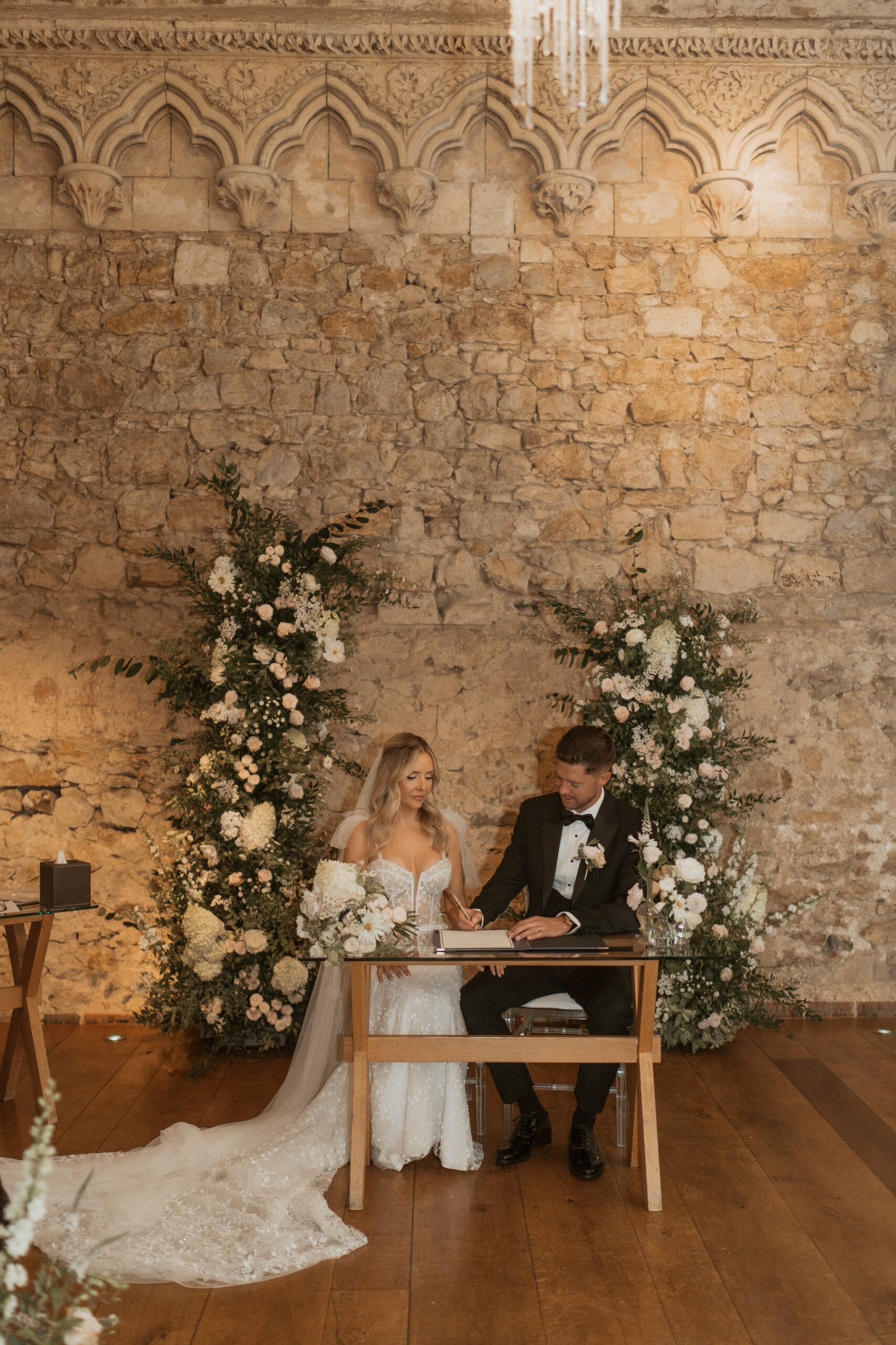 Bride and groom sign the register by floral pillars and carved stone walls in the Monks' Refectory at Notley Abbey
